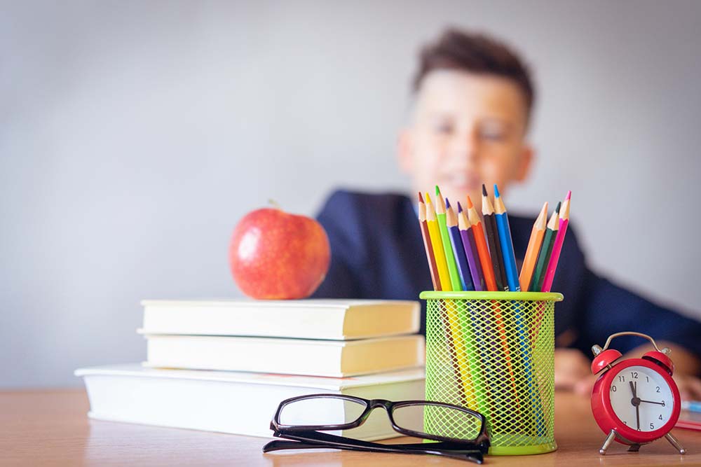 A child next to a teacher’s desk with books and an apple